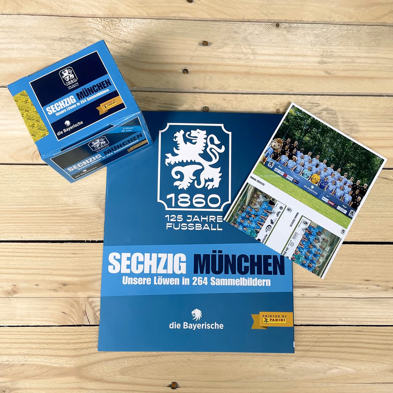 Sammelalbum und Stickerboxen der Edition ‚Sechzig München – Unsere Löwen in 264 Sammelbildern‘ zum 125-jährigen Jubiläum des TSV 1860 München, auf hellem Holzuntergrund.